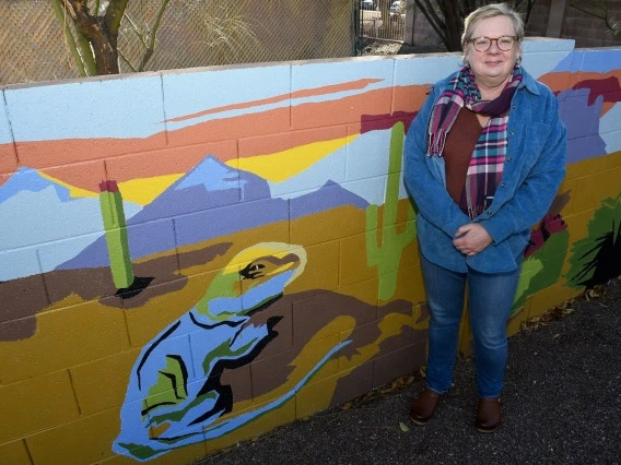 A woman stands in front of a multi-colored mural depicting the desert.